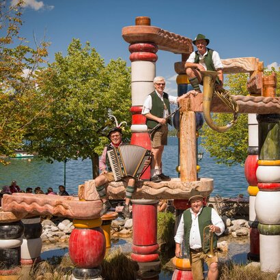Vor dem See stehen vier Männer in Tracht auf einer dekorativen Holzskulptur am Ufer des Zeller Sees.