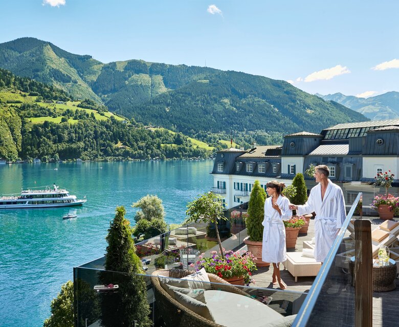 Ein Paar im Bademantel steht auf der Hotelterrasse mit Blick über den See und die Berge.
