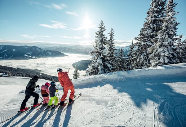 Vier Personen stehen mit Skiern an einer verschneiten Piste und blicken über die winterliche Seenlandschaft.