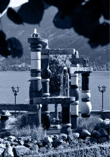 Eine skulpturale Brunnenanlage steht am Ufer des Zeller Sees vor Bergkulisse.