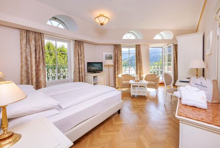 Ein helles Hotelzimmer mit Doppelbett, Sitzbereich und offenem Balkon mit Blick auf den See.