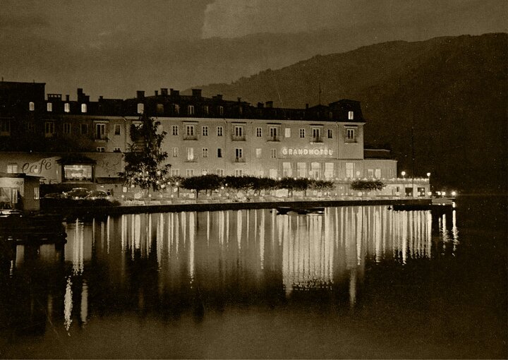 Ein historisches Hotelgebäude spiegelt sich nachts ruhig im Wasser des Zeller Sees.