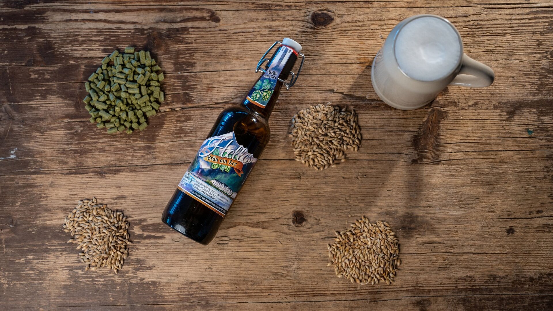 A beer bottle lies on a rustic wooden table, surrounded by malt and hops, with a filled mug on the right.