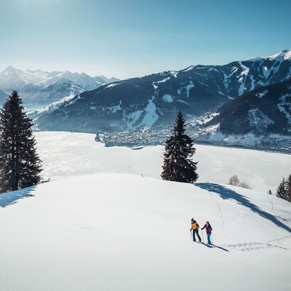 Two winter sports enthusiasts walk across a snowy hillside overlooking Lake Zell and the surrounding mountains.