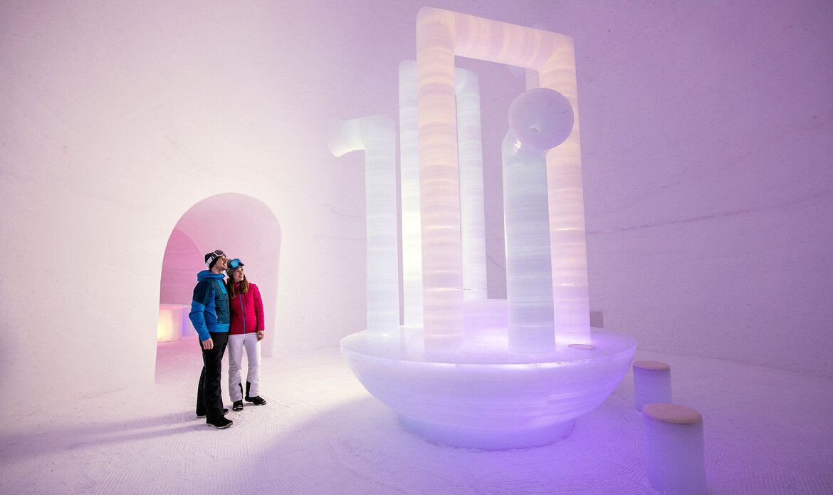 Two people stand inside an artfully lit ice installation with an arched opening and rounded forms.