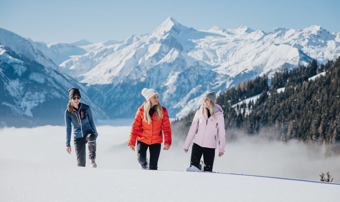 Three women are walking across a snowy slope with a winter mountain landscape behind them.