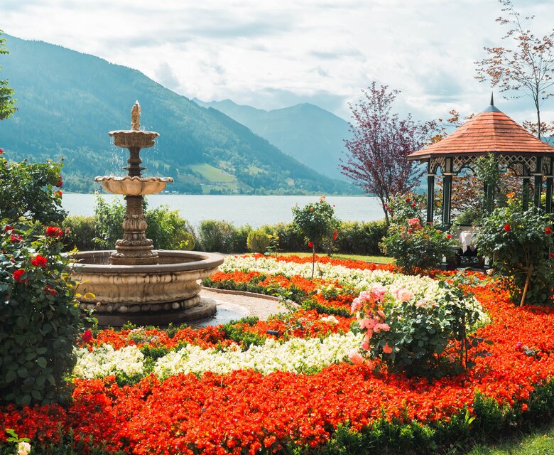 Ein farbenfroher Sommergarten mit Brunnen, Blumenbeeten, Pavillon und Blick auf den Zeller See und die Berge.