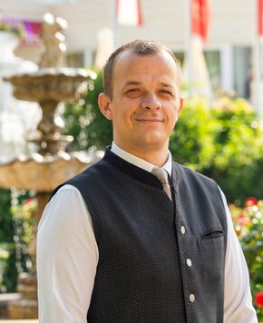 Ein Gastgeber steht vor einem Brunnen im Garten des GRAND HOTEL Zell am See.