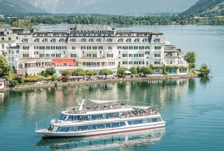 GRAND HOTEL Zell am See am Zeller See Das GRAND HOTEL Zell am See liegt direkt am Seeufer, davor fährt ein Ausflugsschiff über das ruhige Wasser.