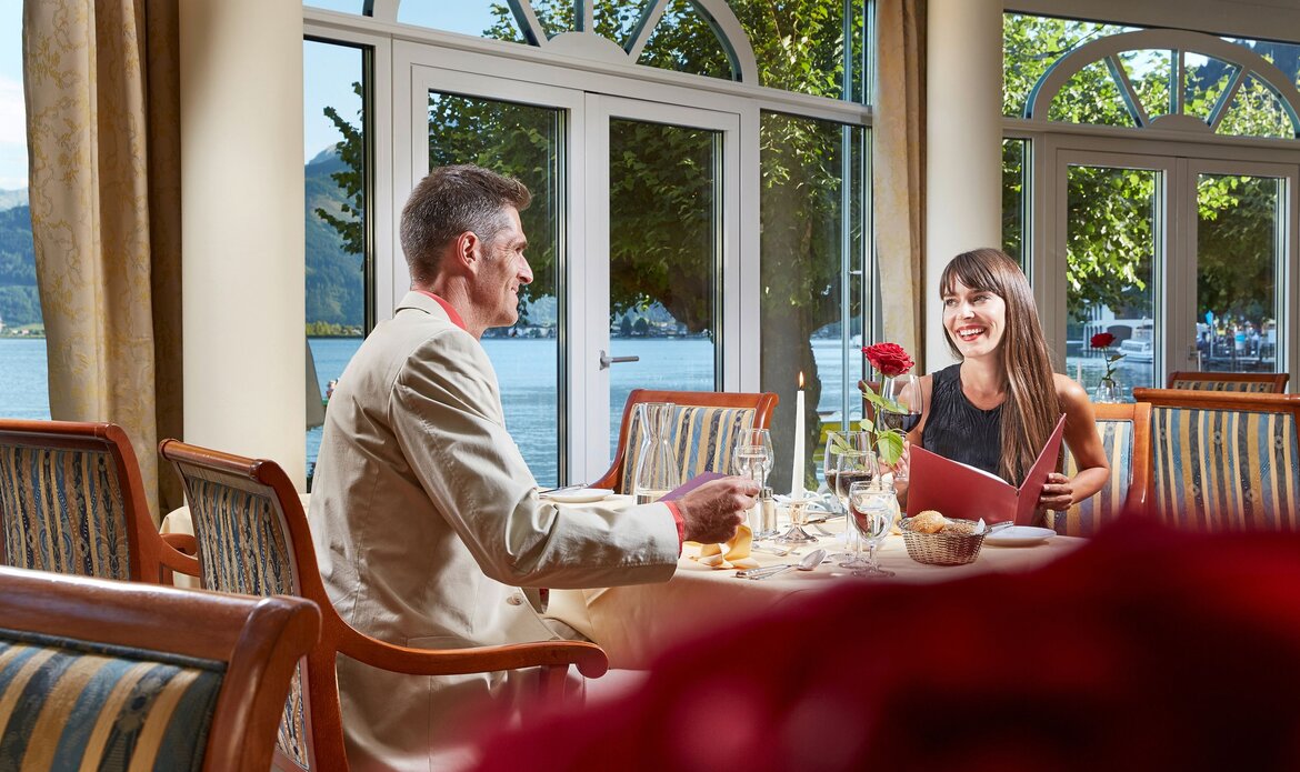 A man and a woman are seated at a table in Restaurant Kaisersaal with a view of Lake Zell through the windows.