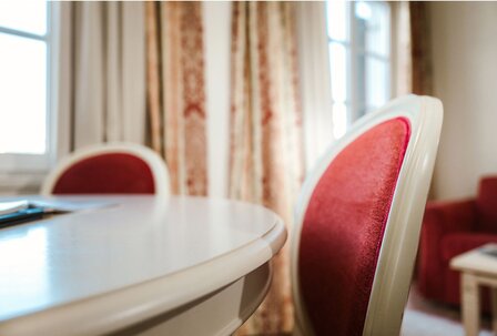 A red upholstered chair stands by a light table in a hotel room with windows and curtains.