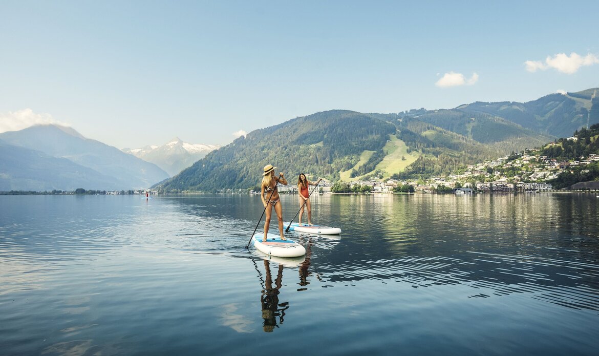 Two people stand on paddleboards on the calm water of Lake Zell with the mountain landscape behind them.