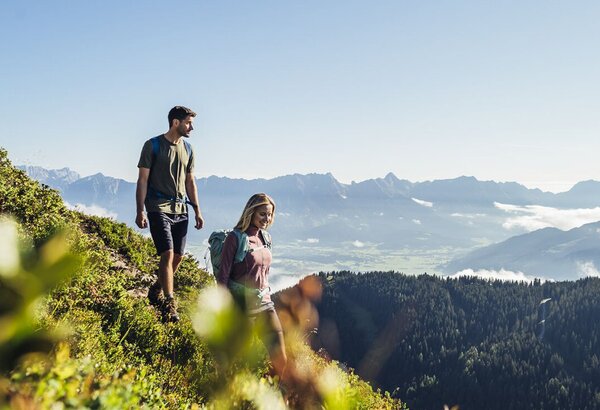 Ein Wanderer und eine Wanderin stehen auf einem bewachsenen Hang mit weitem Blick auf die Berge.
