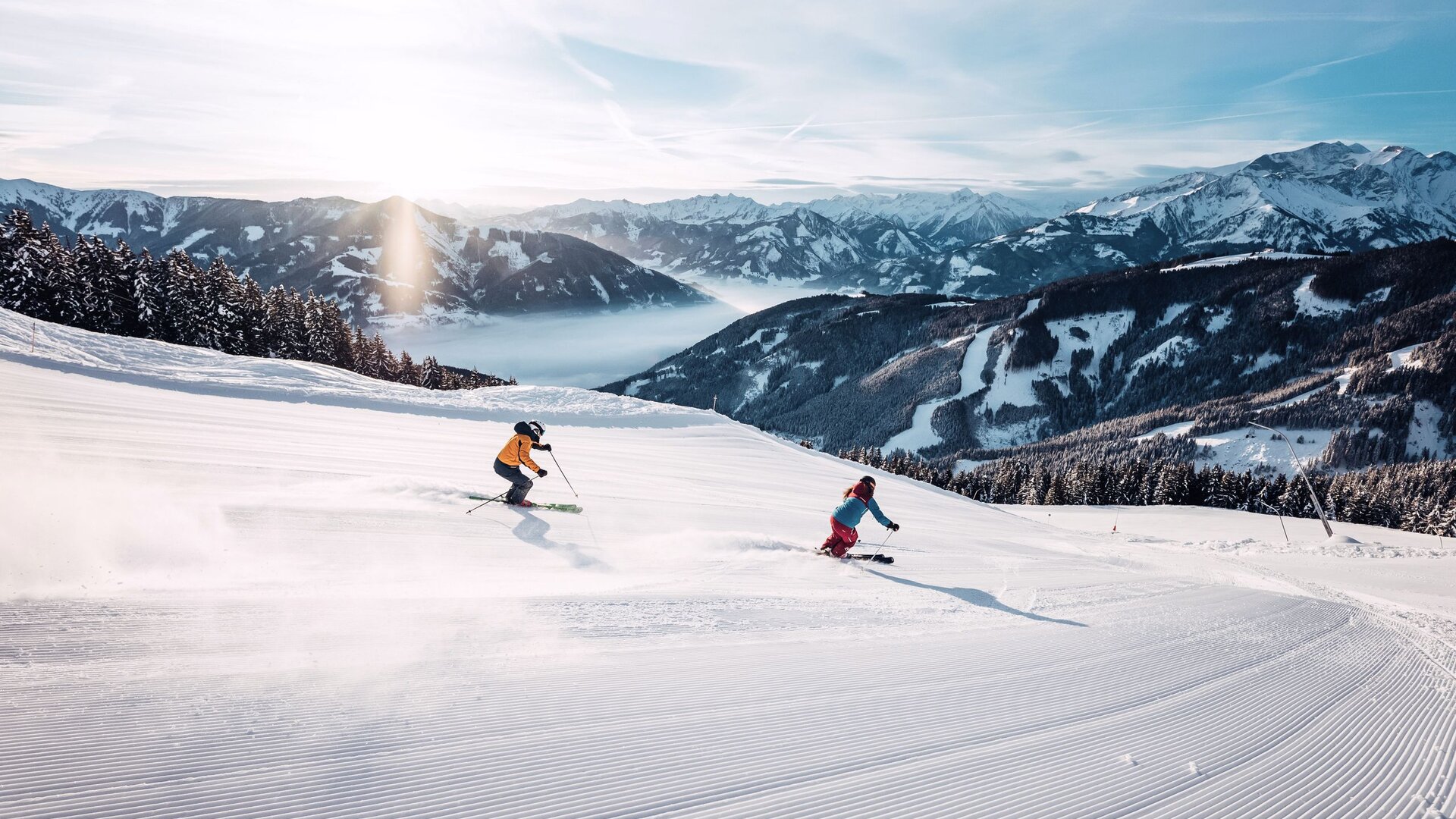 Two skiers descend a wide slope with snowy mountains in the background near Lake Zell.