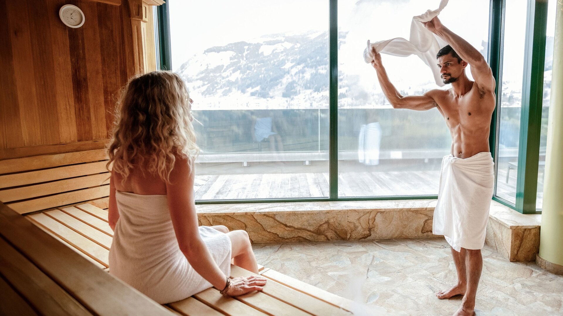 Eine Frau sitzt auf der Holzbank im Ruheraum der Sauna, während ein Mann sich im Handtuch abtrocknet.