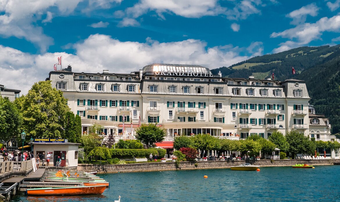 Das Grand Hotel liegt direkt am Zeller See, davor sind Stege, Boote und Spaziergänger am Ufer zu sehen.