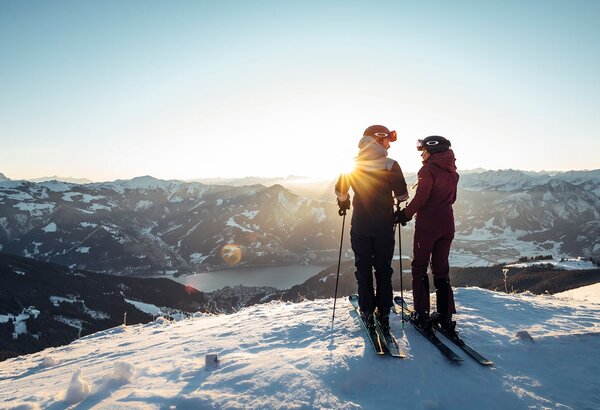 Two winter sports enthusiasts stand on a snowy ridge and look out over Lake Zell and the valley below.