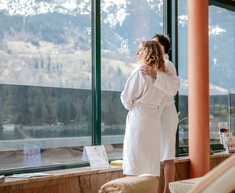 Zwei Frauen im Bademantel stehen vor großen Fenstern im Ruheraum mit Blick auf die Berge und den See.