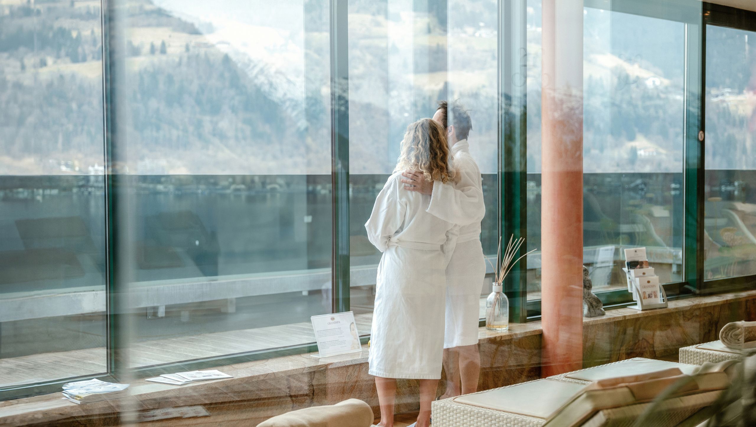 Eine Frau im Bademantel steht vor der Glasfront im Wellnessbereich mit Blick auf die Berge.
